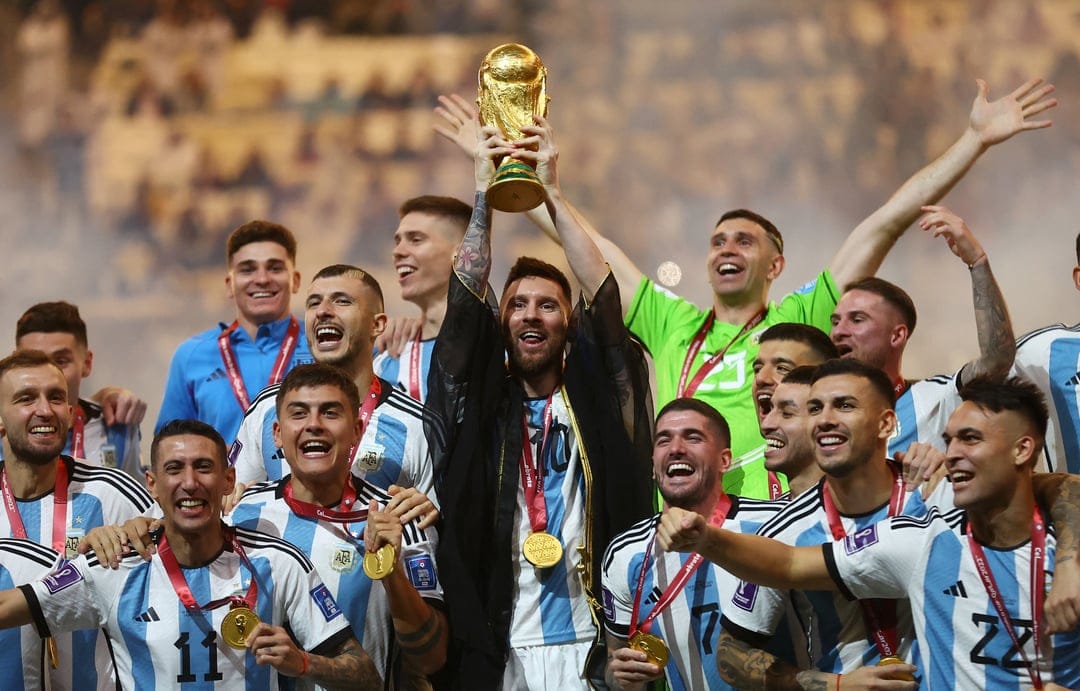 Lionel Messi hoists the gold FIFA World Cup trophy while surrounded by teammates in blue and white jerseys.