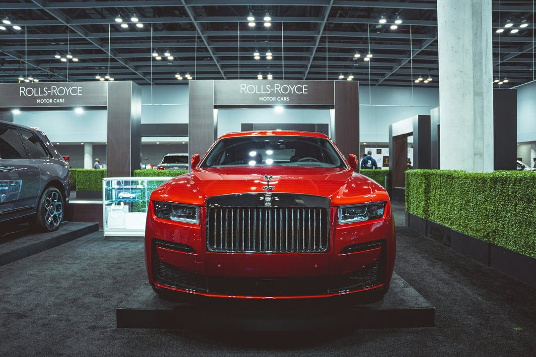 Red Rolls-Royce sedan on a platform in front of a green hedge wall and luxury branding at an auto show booth.
