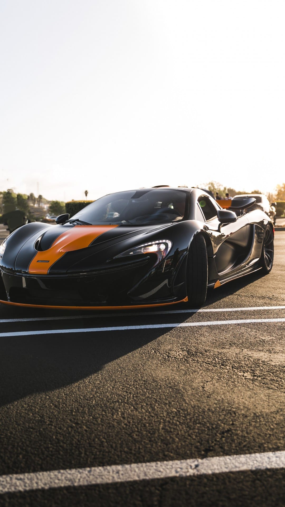 Black McLaren P1 supercar with orange stripes parked on asphalt during a warm golden hour sunset.