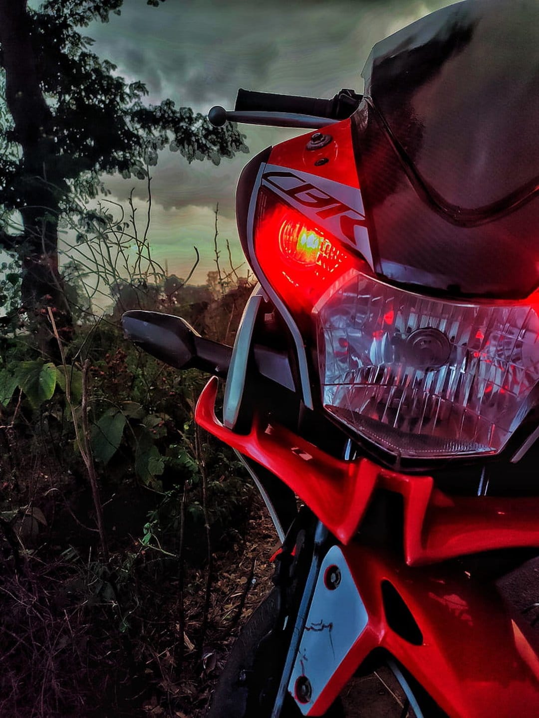 Red motorcycle headlight glowing with a warm beam against a purple and orange sunset sky with dark clouds.