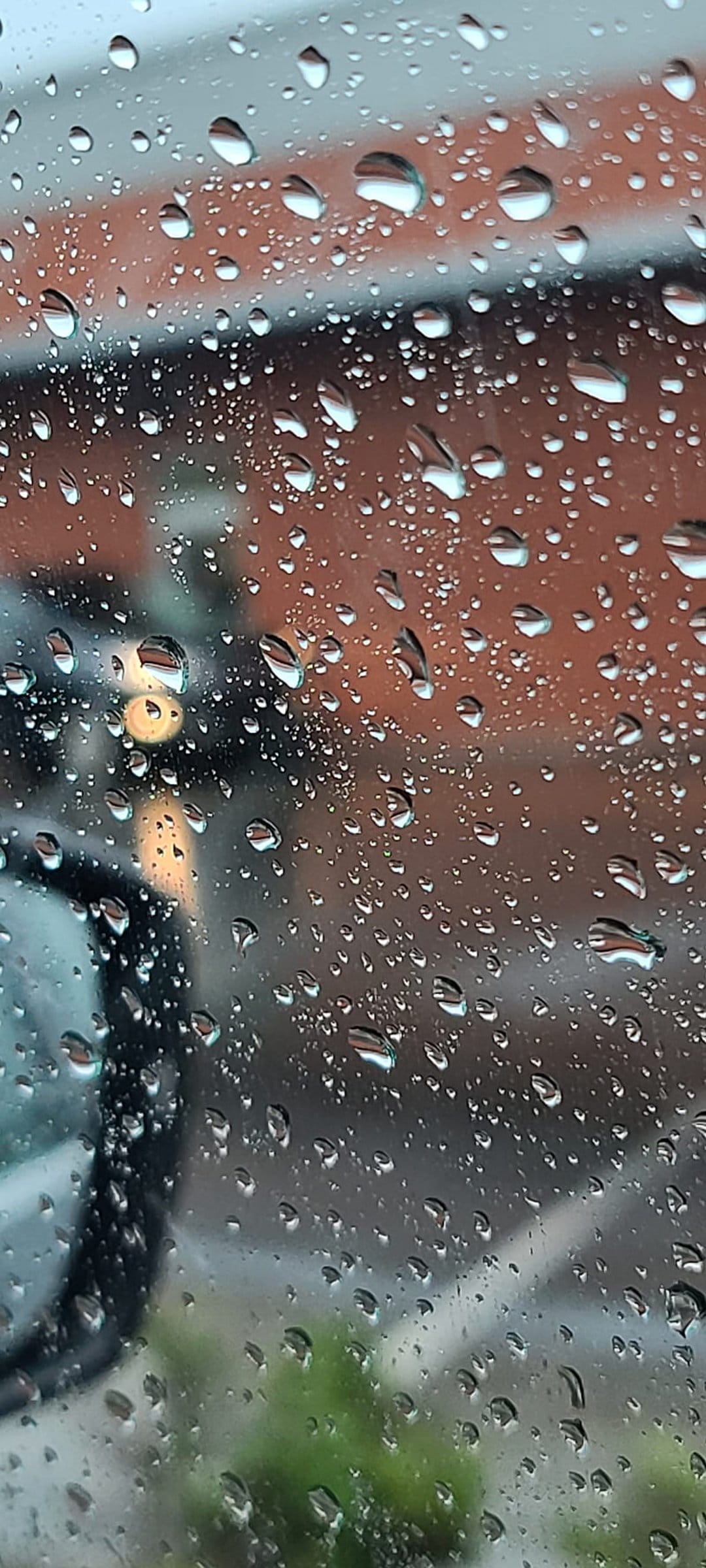 Raindrops on clear glass distort a blurred red brick wall and green leaves in a vertical macro composition.