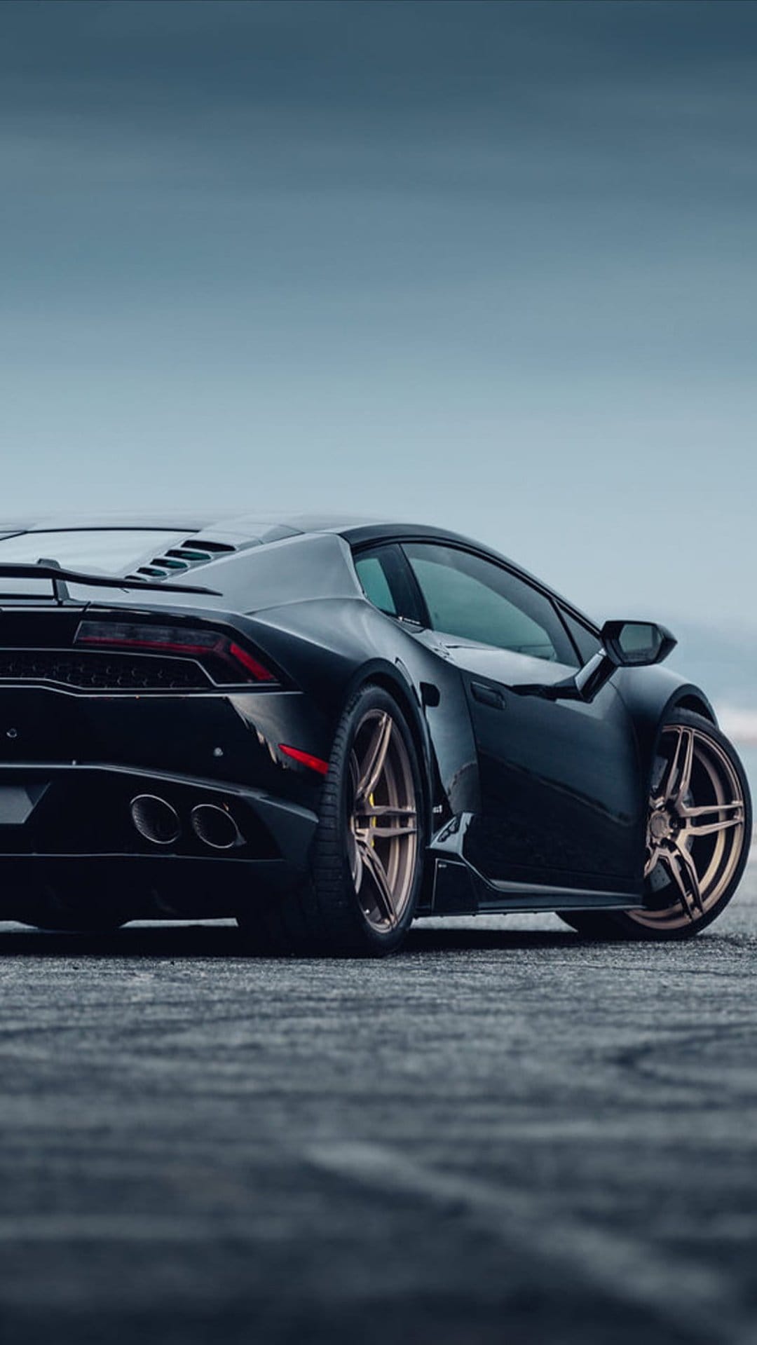 Black Lamborghini supercar with gold rims parked on dark pavement under a cloudy sky viewed from the rear.