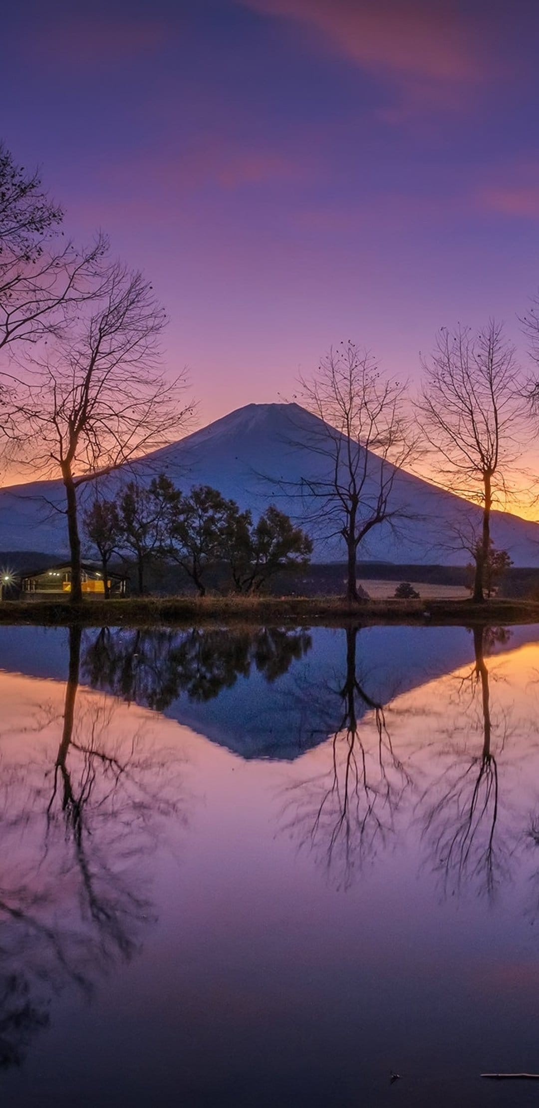 Snow-capped Mount Fuji reflects in a still lake framed by dark trees under a purple and orange sunset sky.