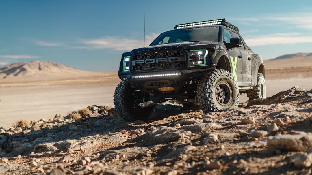 Black Ford Raptor truck with green headlights on rocky desert ground under a bright clear blue sky.