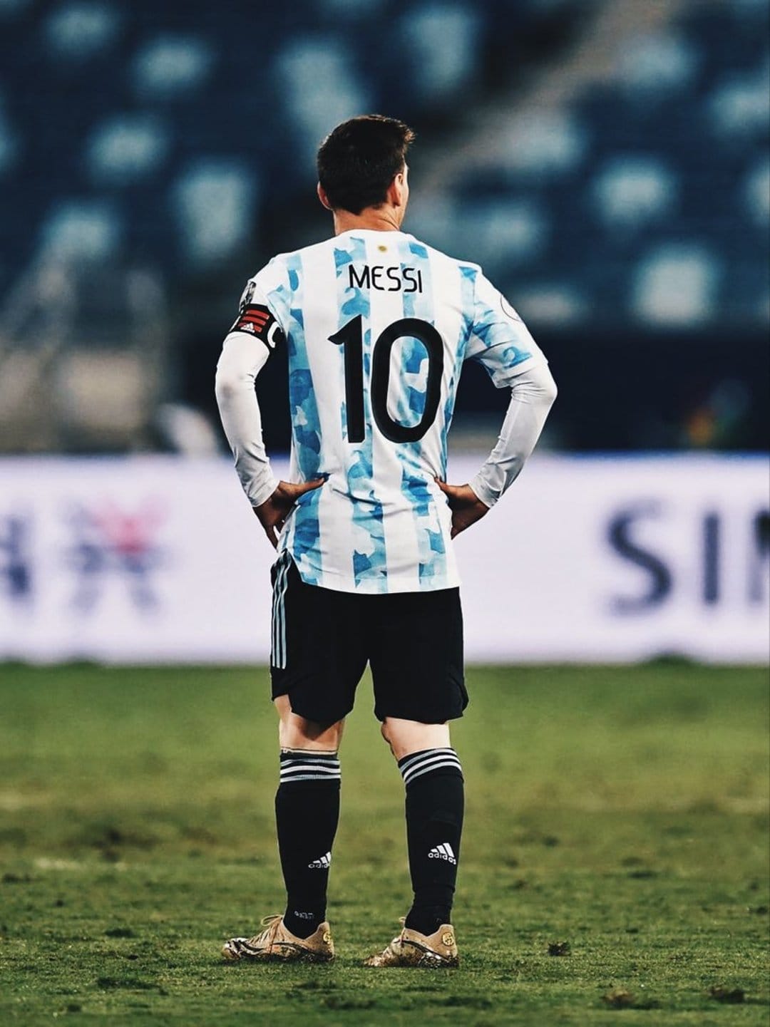 Lionel Messi stands from behind wearing a number 10 Argentina jersey on a green football pitch in a stadium.