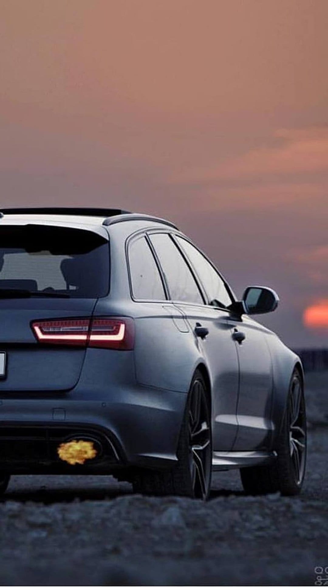 Dark grey Audi RS6 Avant wagon with orange exhaust flames and red taillights against an orange sunset sky.