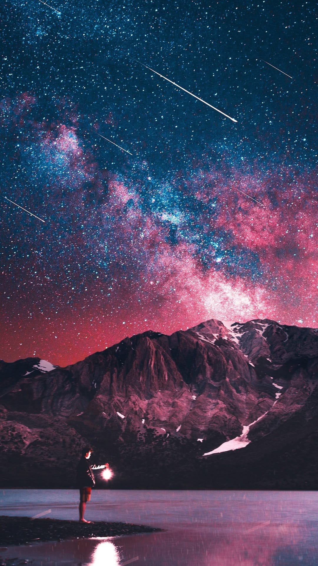 A person holding a glowing lantern stands beside a calm lake reflecting a pink and blue star-filled night sky.