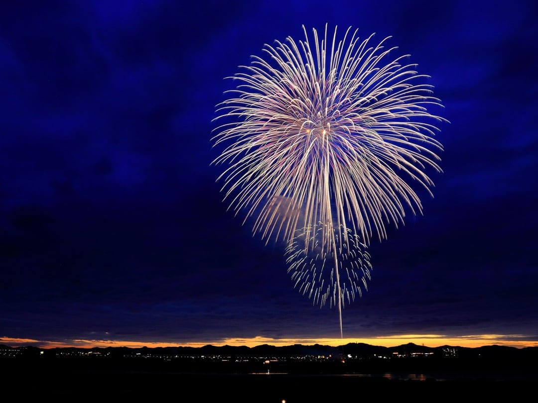 Gold and white fireworks explode over a dark landscape silhouette and a hazy orange horizon in a night sky.