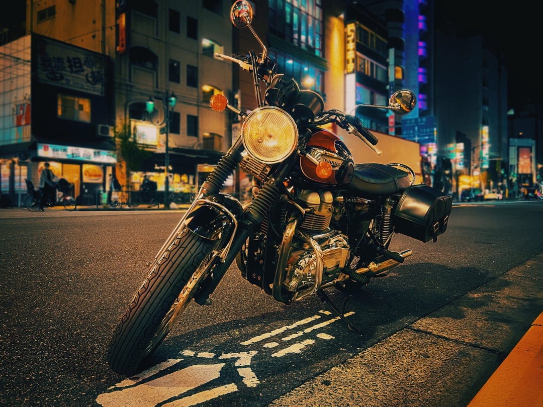 Classic motorcycle with a glowing headlight parked on a wet street reflecting vibrant neon city signs at night.