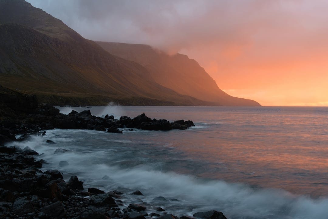 Misty Icelandic mountains and crashing surf under a vibrant orange sunset sky with dark volcanic coastal rocks.