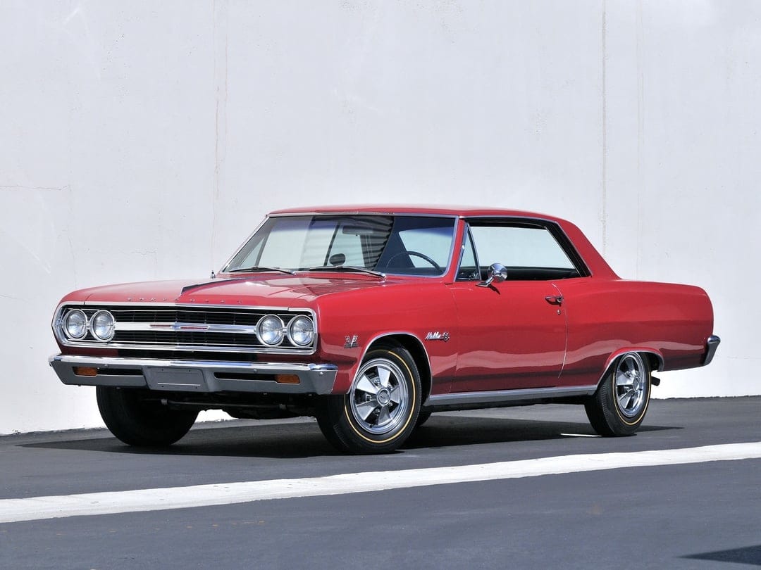 Red 1965 Chevrolet Chevelle Malibu SS classic muscle car with chrome trim on asphalt against a white wall.