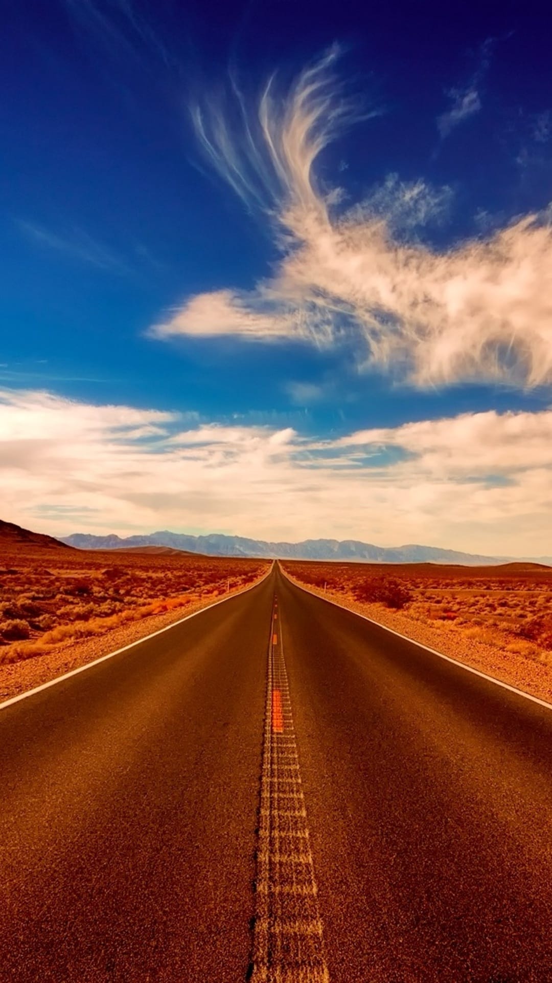 Asphalt road stretches through a vast sun-baked desert toward distant mountains under a bright blue sky with clouds.