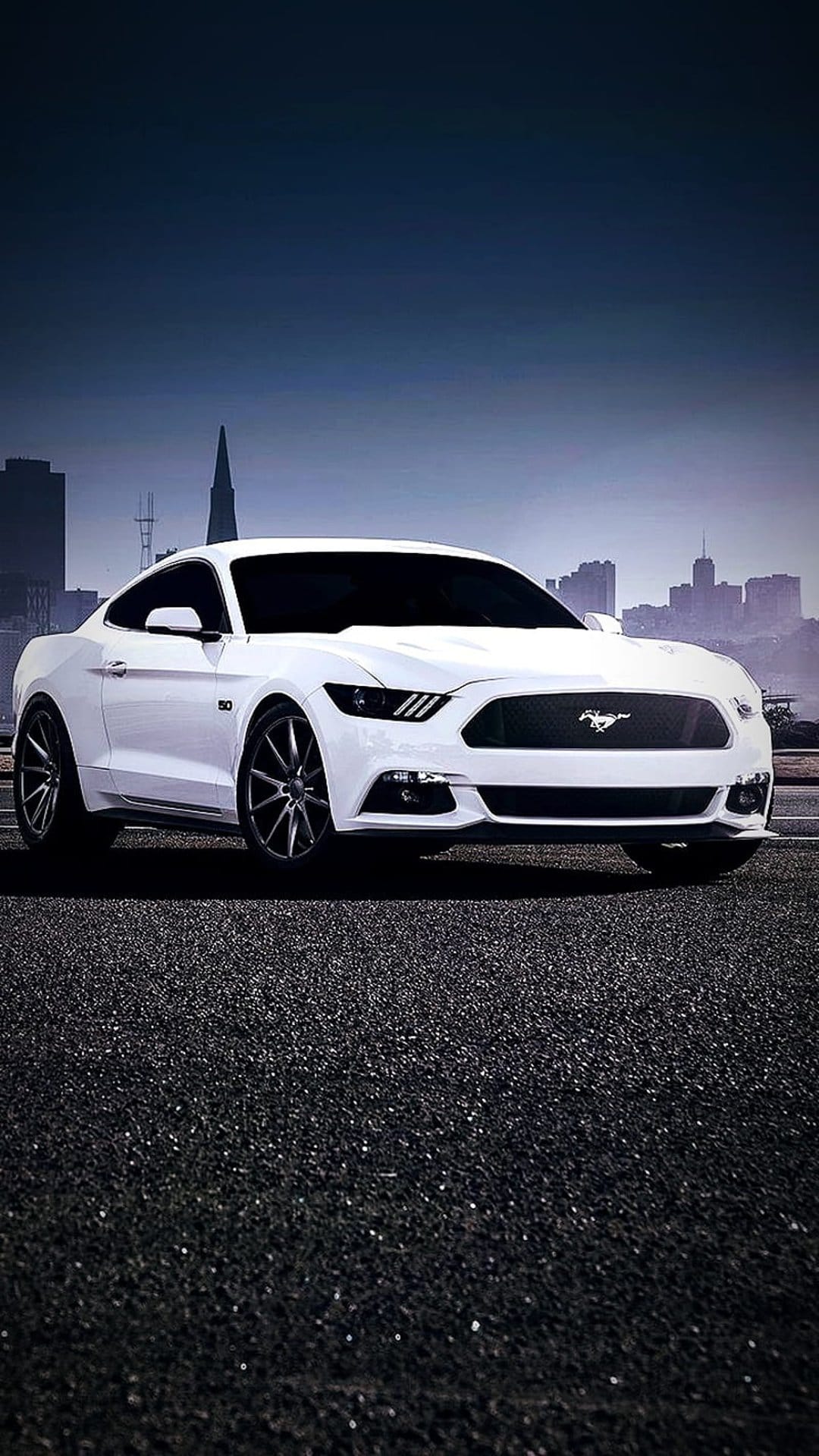White Ford Mustang coupe parked on dark textured asphalt with a blurred cityscape and deep blue sky behind it.