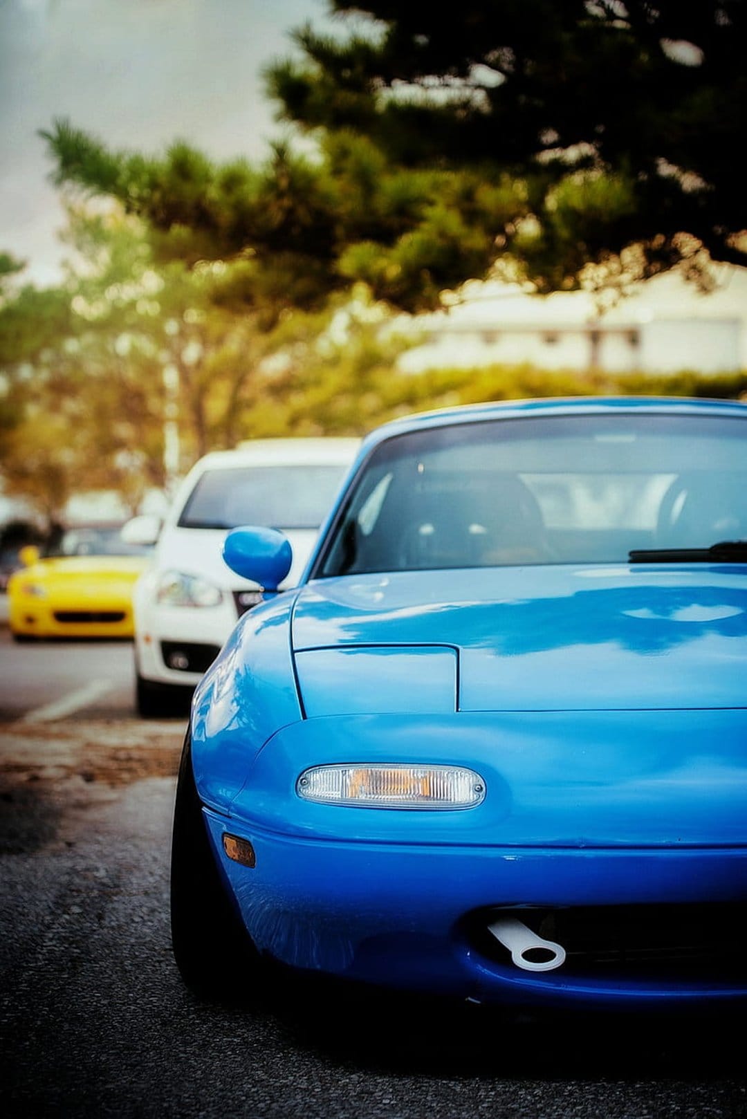 Blue Mazda Miata with pop-up headlights sits parked in a row near a yellow sports car and lush green trees.