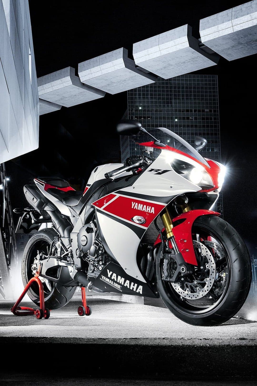 Yamaha R1 sportbike on a red stand at night with glowing headlights against a dark urban architecture backdrop.