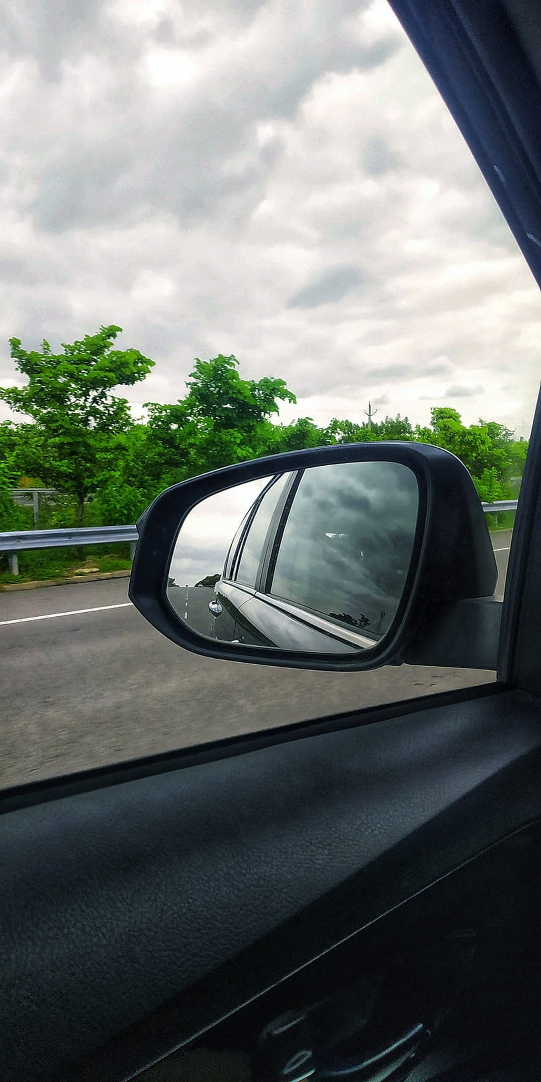 Car side mirror reflecting a green forest and highway under a cloudy sky with the car window visible inside.