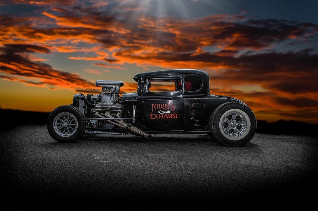 Black custom hot rod with a chrome engine and side exhaust against a dramatic orange sunset sky.