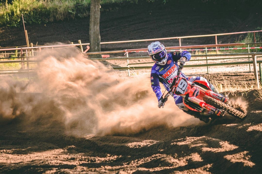 Motocross racer on bike number 19 leans into a turn, kicking up a large cloud of brown dust on a dirt track.