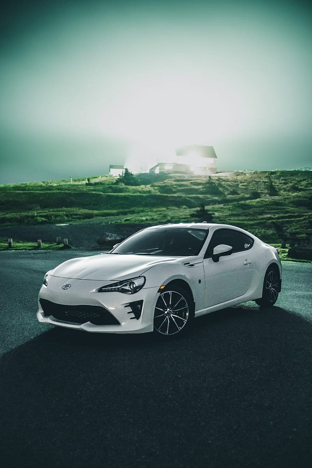 Toyota 86 sports car parked on asphalt with thick fog, rolling green hills, and a teal sky background.