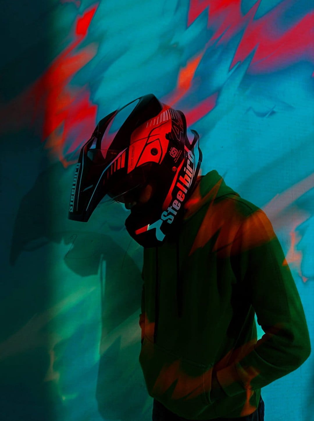 Motocross rider in a black helmet and green hoodie stands against a wall with red and teal neon lighting.