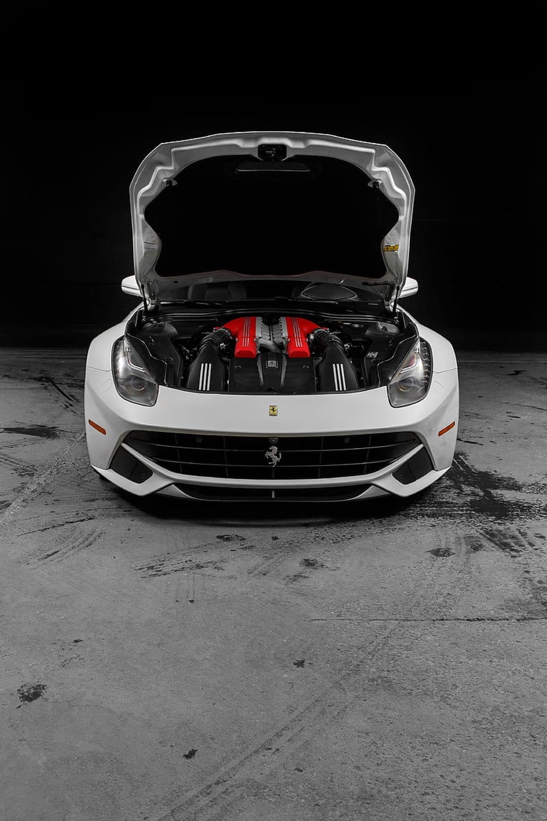 White Ferrari F12 with an open hood reveals vibrant red engine covers against a dark minimalist background.