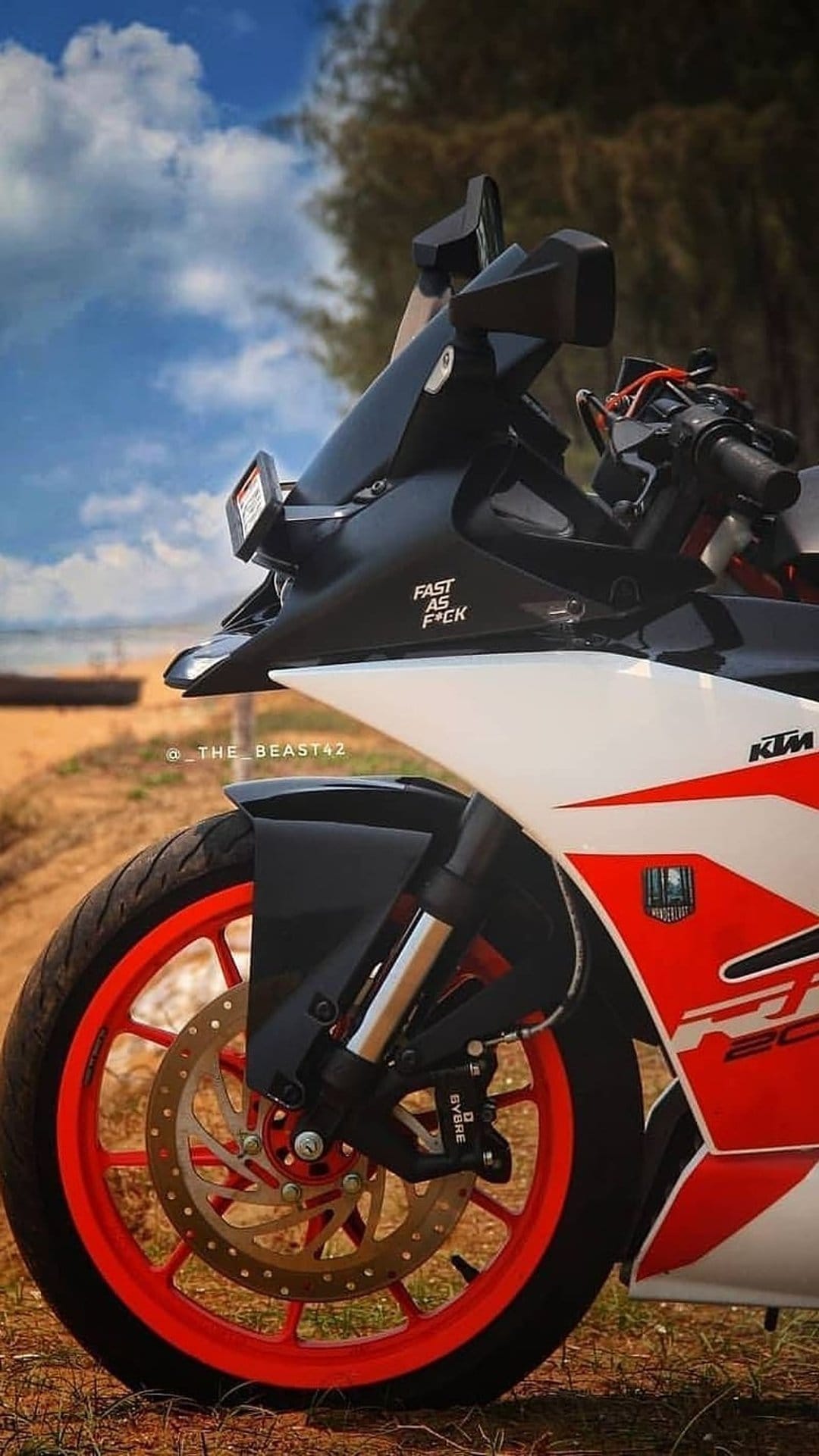 KTM motorcycle front wheel and orange rim stand near a coastal beach under a bright blue sky with white clouds.
