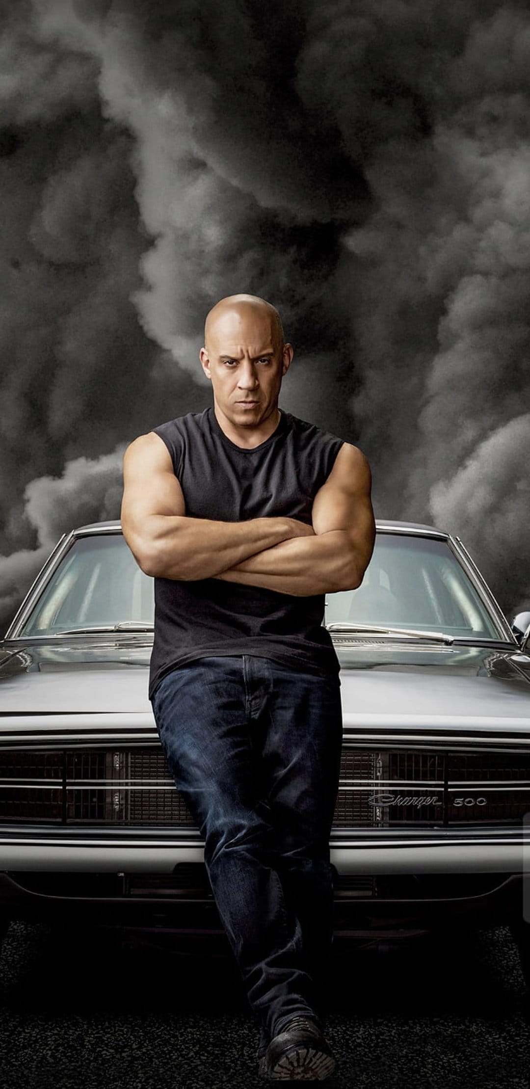 Vin Diesel stands with arms crossed in front of a black Dodge Charger surrounded by dense gray smoke clouds.