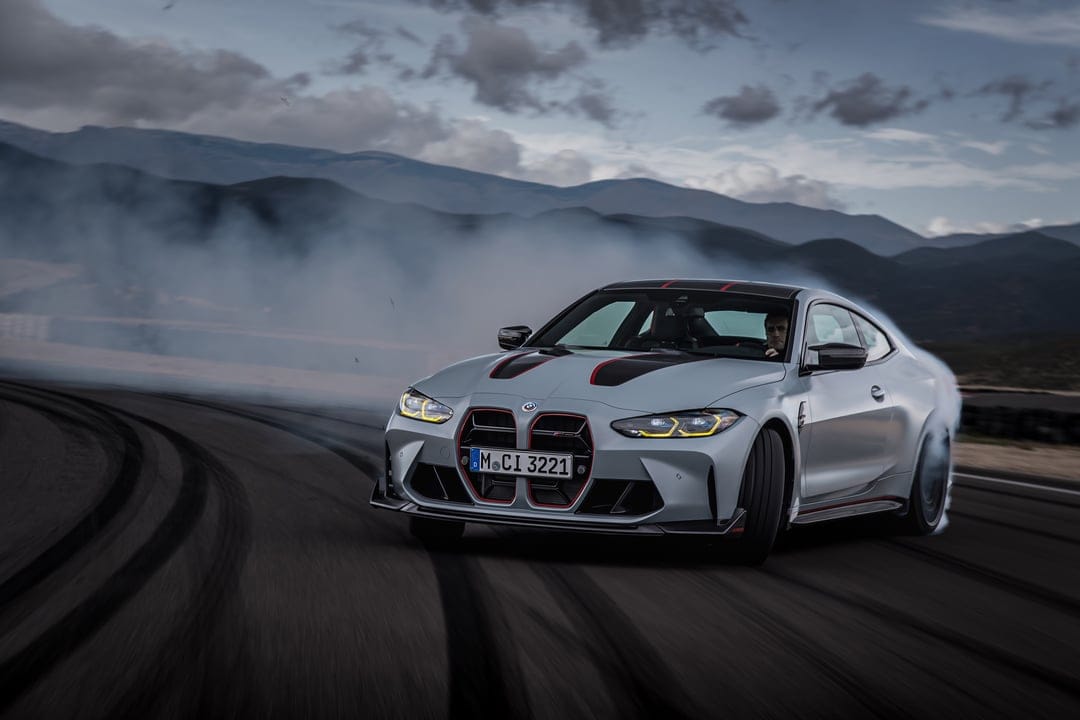 Silver BMW M4 CSL sports car drifting on a dark track with thick white smoke against a mountain backdrop.
