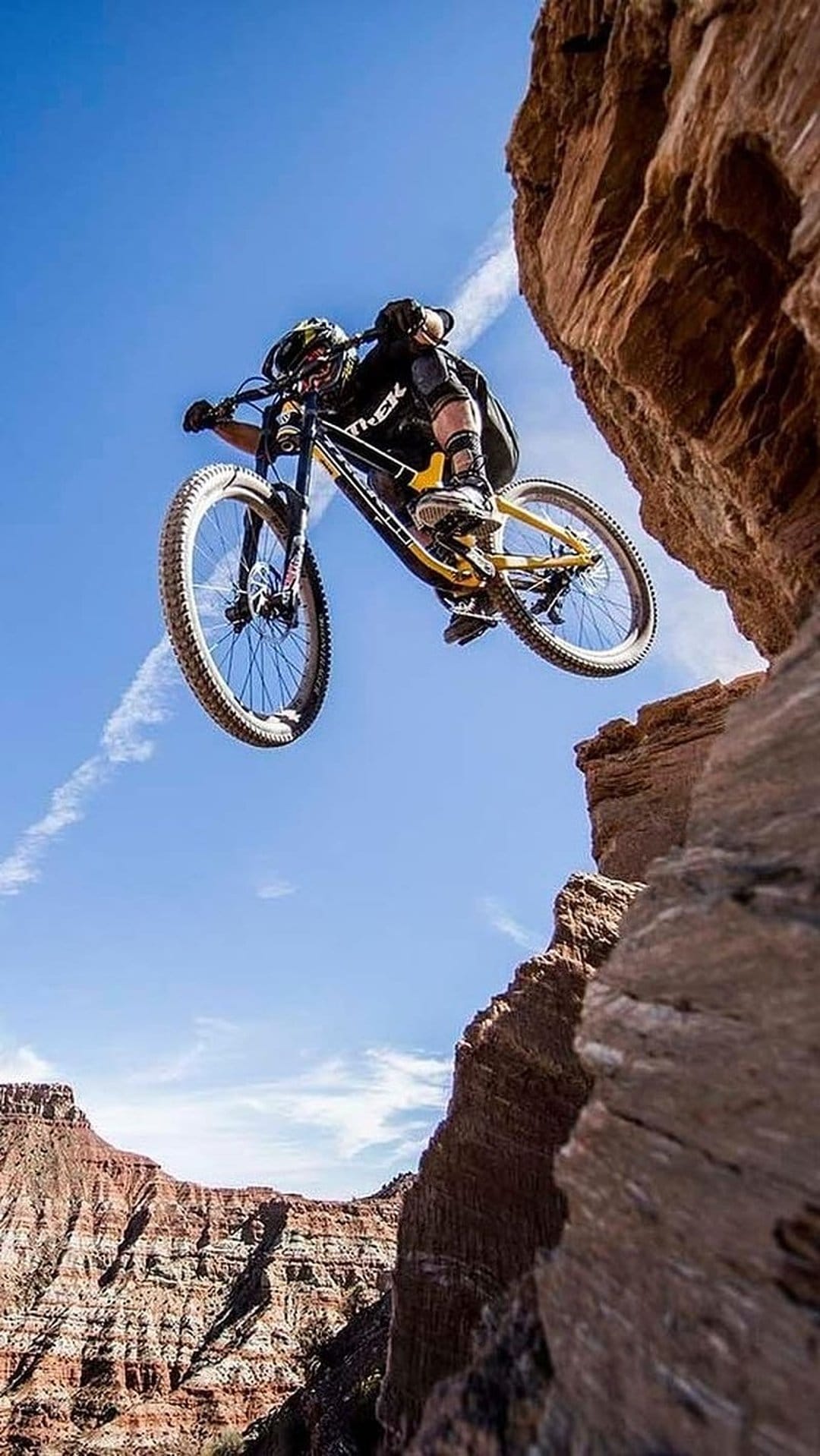 Mountain biker in mid-air jumping off a rocky desert cliff against a clear blue sky with thin wispy clouds.