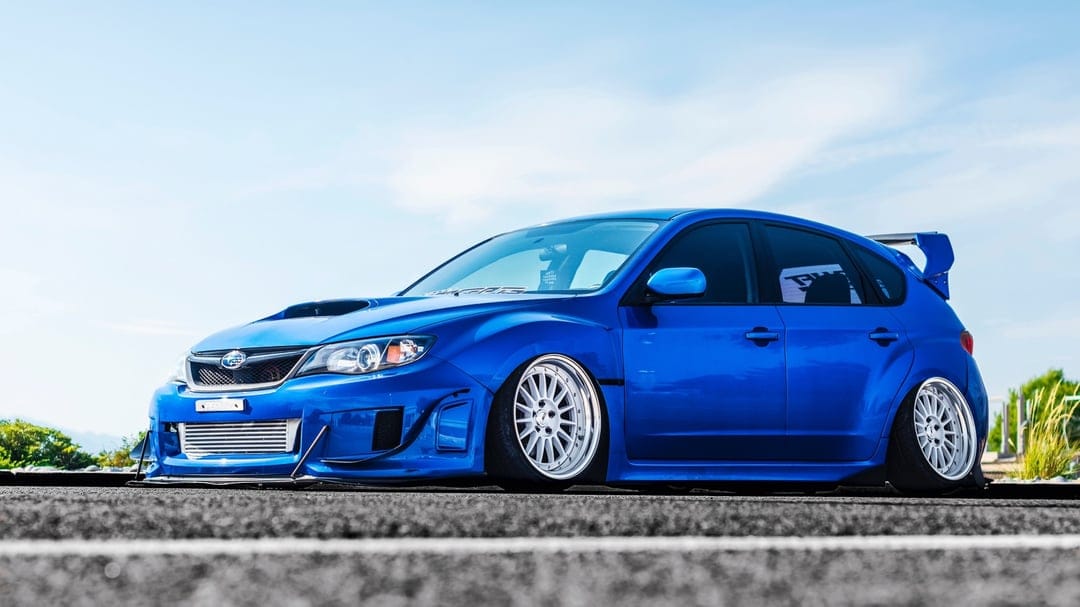Low-angle view of a slammed blue Subaru WRX STI hatchback with white deep-dish wheels on a road under a blue sky.