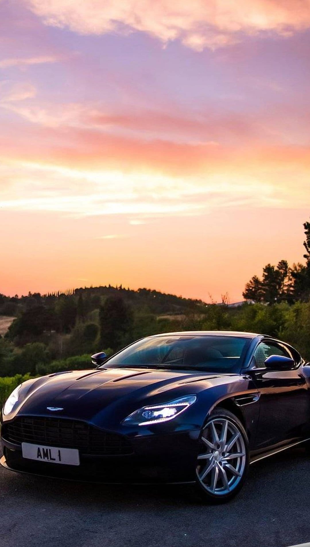Aston Martin DB11 with glowing headlights on a Tuscan road during a vibrant orange and purple sunset.