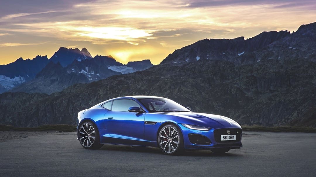 Metallic blue Jaguar F-Type parked on a mountain road with a golden sunset over jagged peaks in the background.