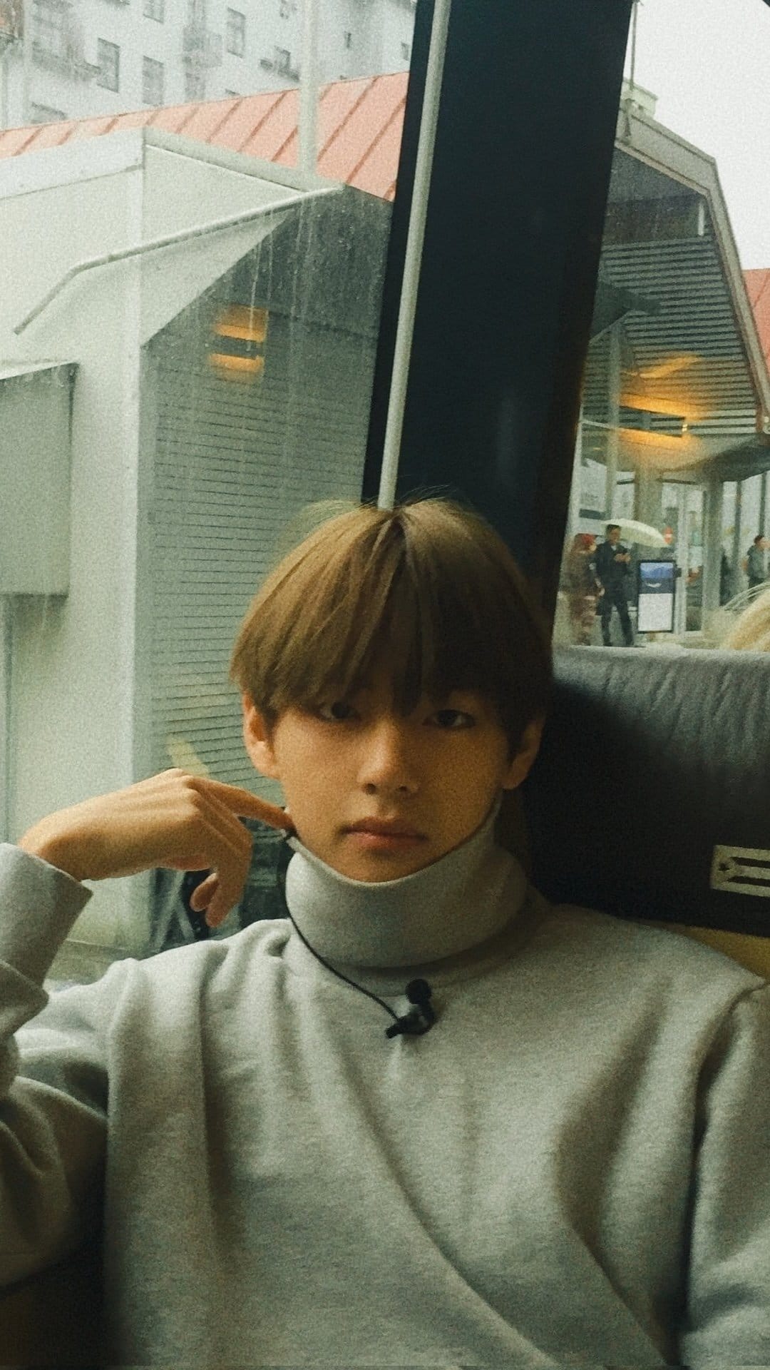 Young man in a grey turtleneck rests his hand on his chin against a rainy window in this close-up portrait.