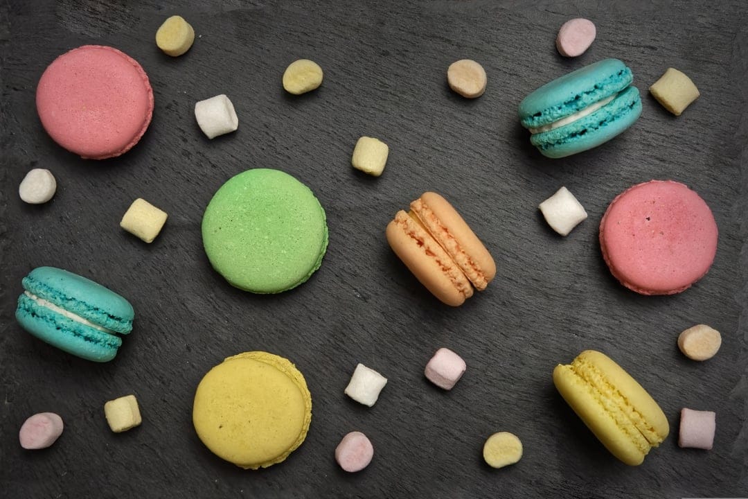 Colorful macarons and mini marshmallows scattered across a dark textured slate background in a flat lay view.