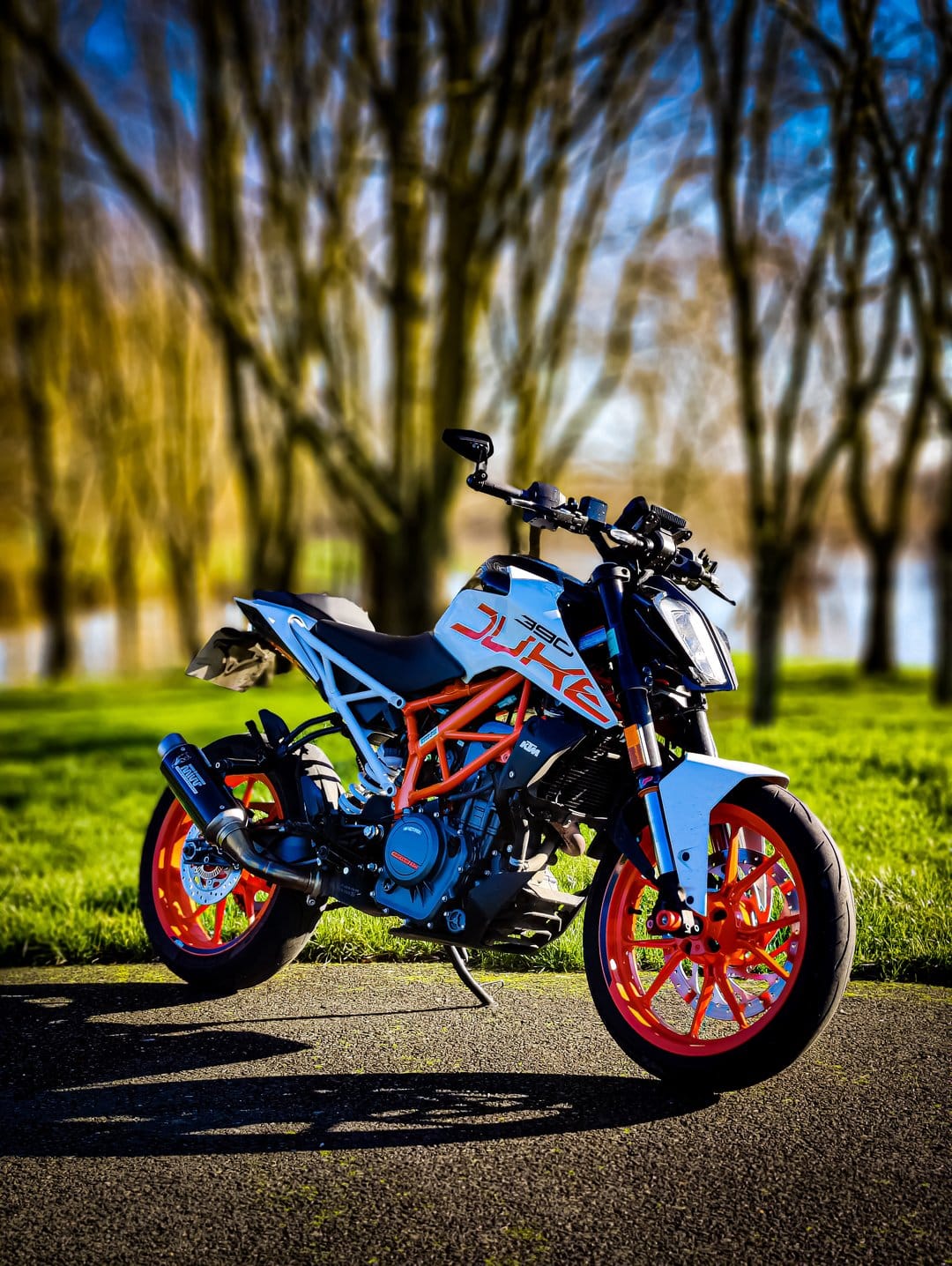 KTM Duke motorcycle with orange wheels parked on asphalt against a blurred green forest background in daylight.