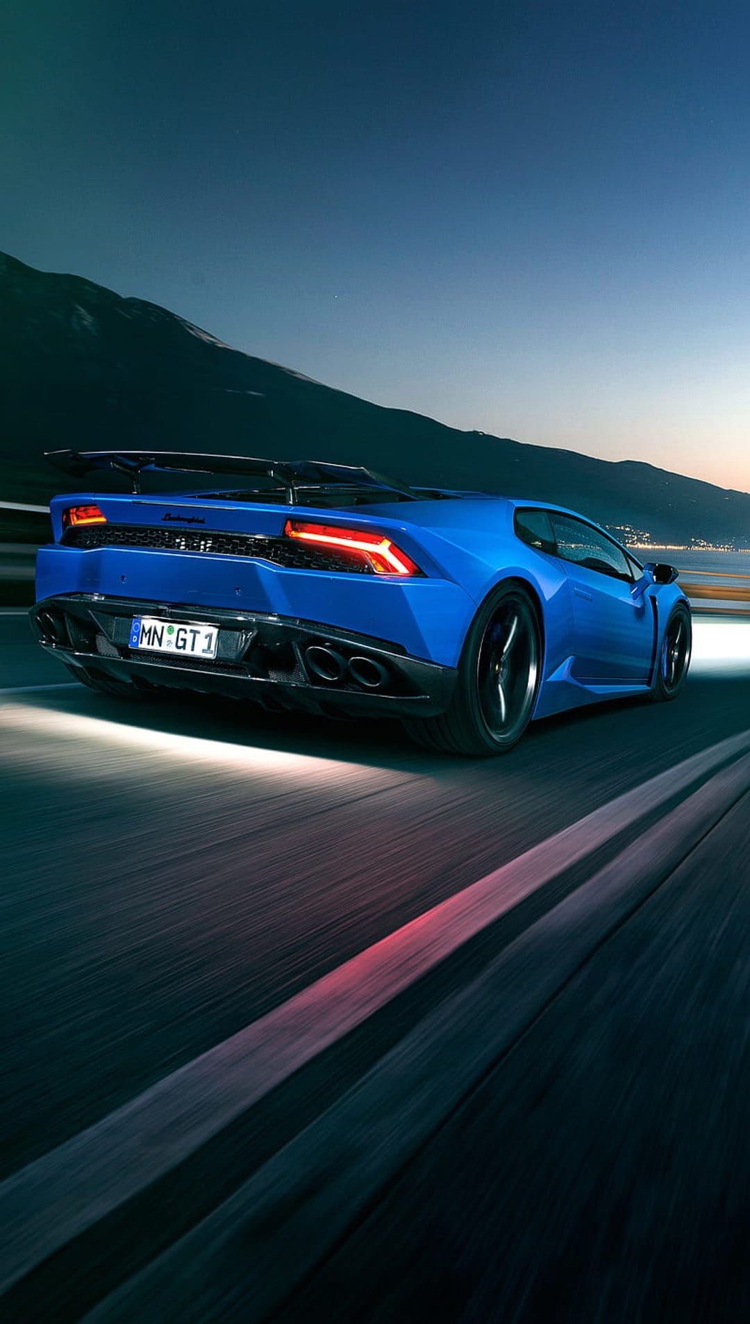 Blue Lamborghini Huracan GT1 driving on a winding mountain road at dusk with glowing red taillights and spoiler.