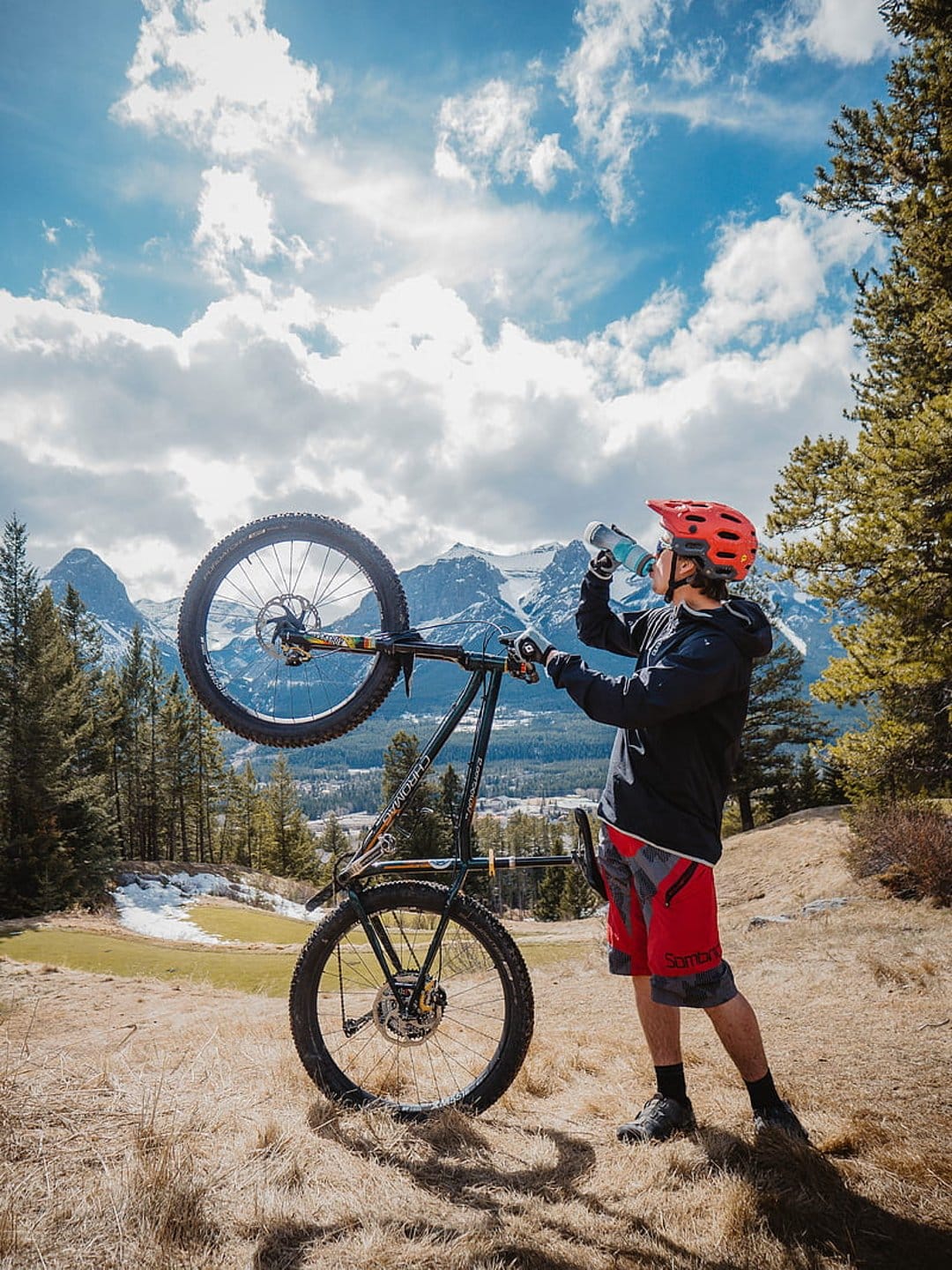 Male cyclist in red shorts drinks from a bottle beside his bike with snow-capped alpine mountains behind him.