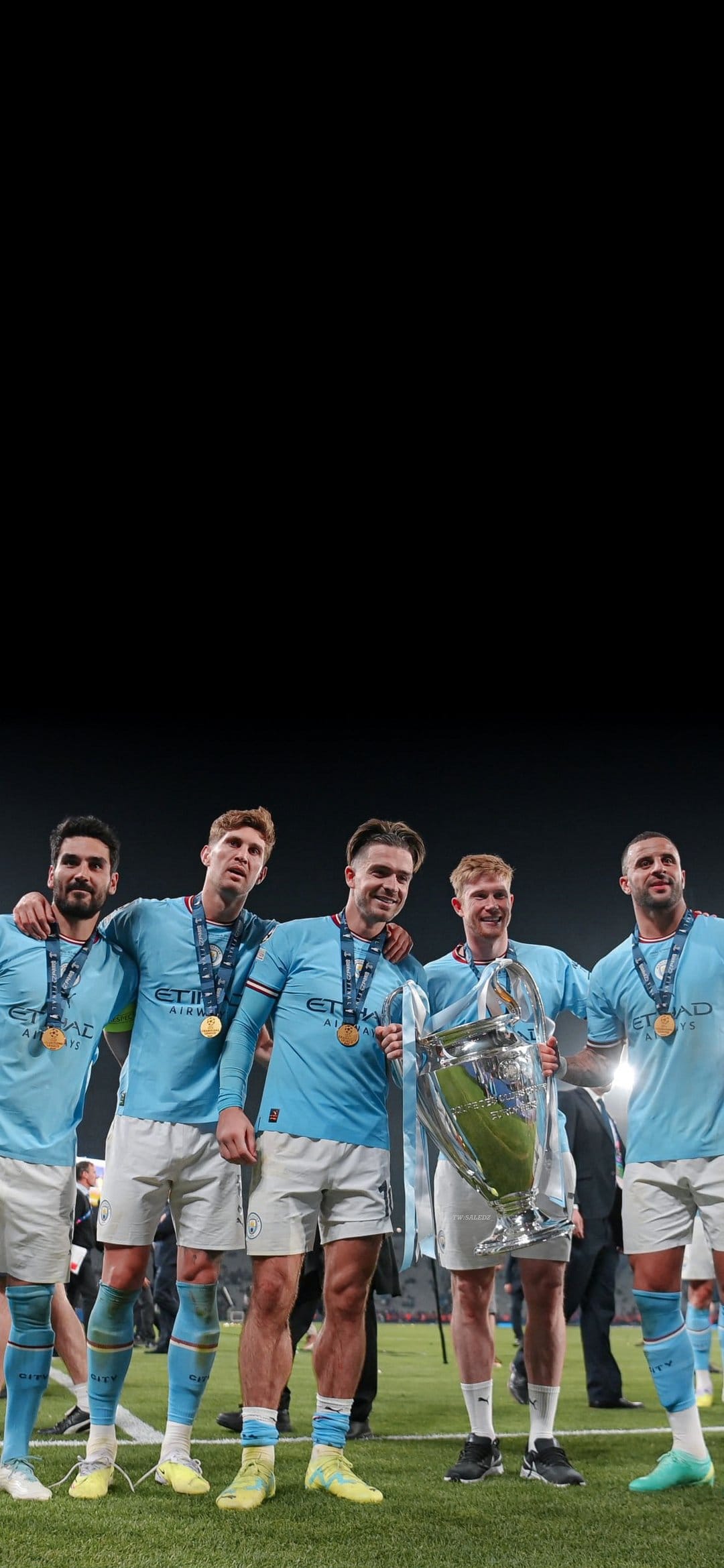 Five Manchester City players hold the silver Champions League trophy while wearing medals on a football pitch.