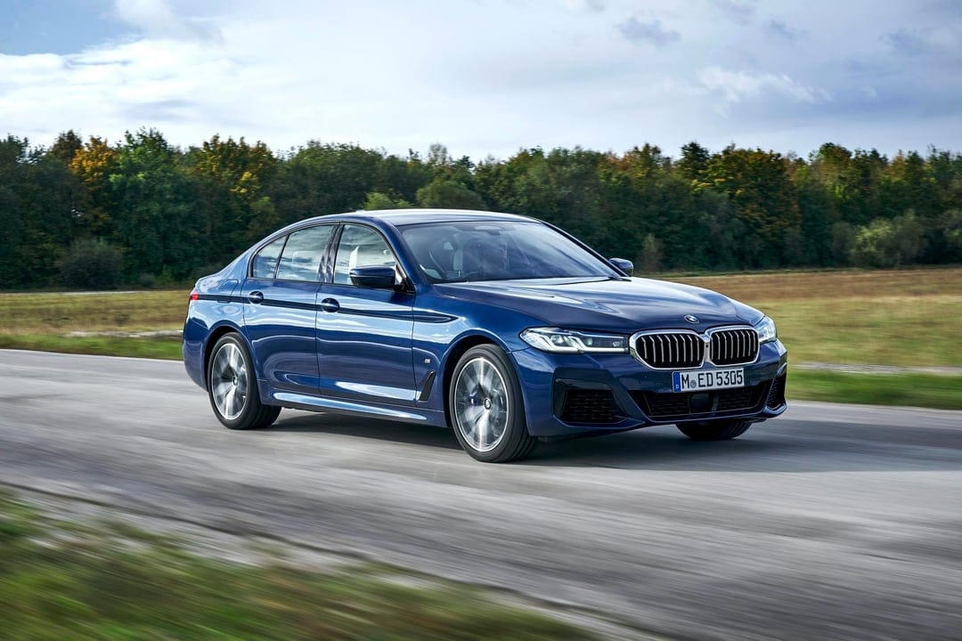 Dark blue BMW sedan driving on a scenic road with motion-blurred wheels and a forest background under a cloudy sky.