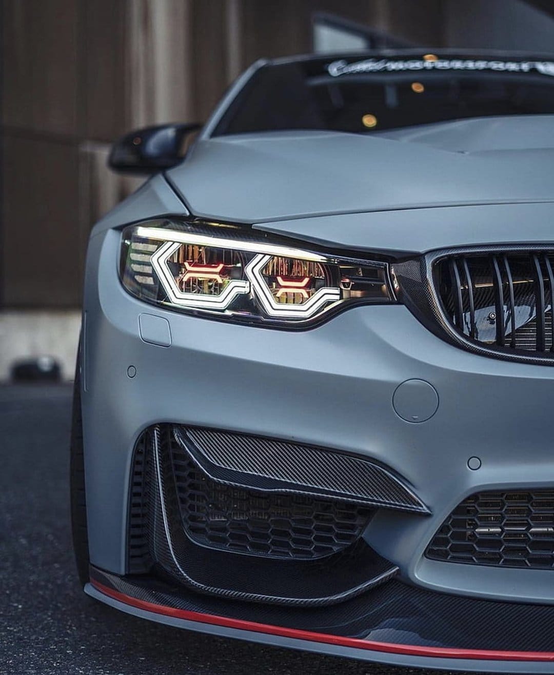 Matte grey BMW M4 front bumper with custom hexagonal LED headlights, carbon fiber accents, and orange light rings.