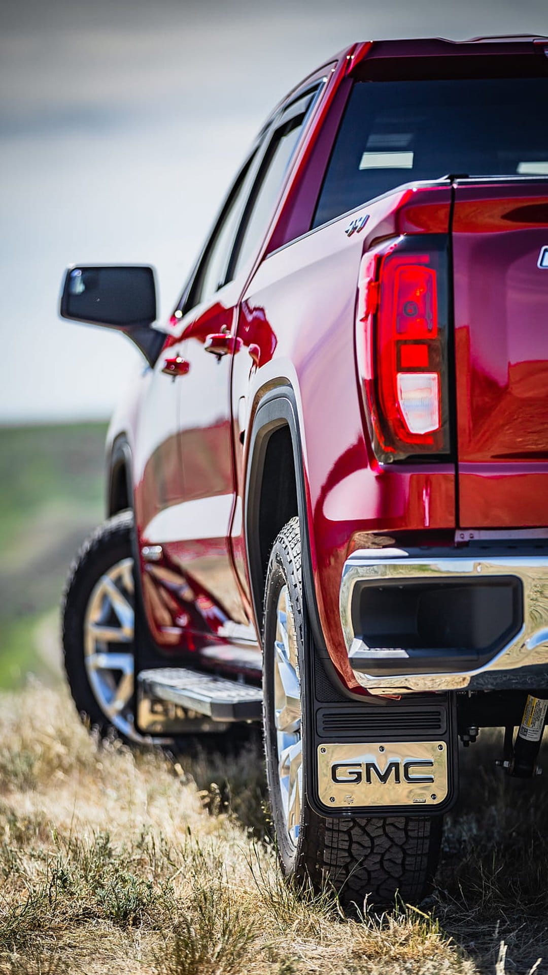 Red GMC truck tailgate and chrome bumper parked on a dry grassy hill under a bright sky with rugged tire treads.