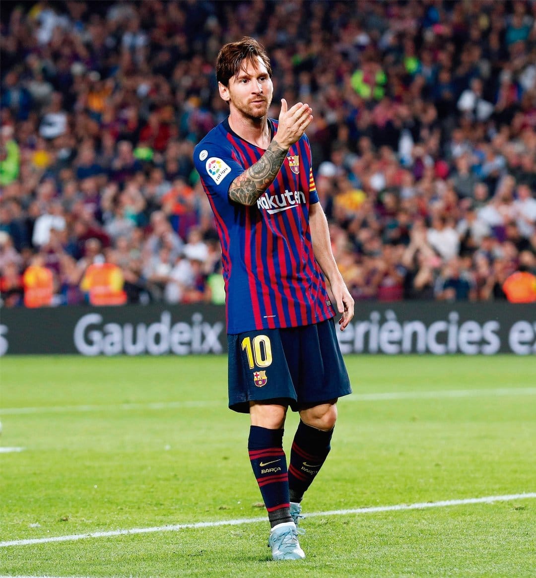 Lionel Messi in a blue and red Barcelona jersey stands on a green football field during a professional match.