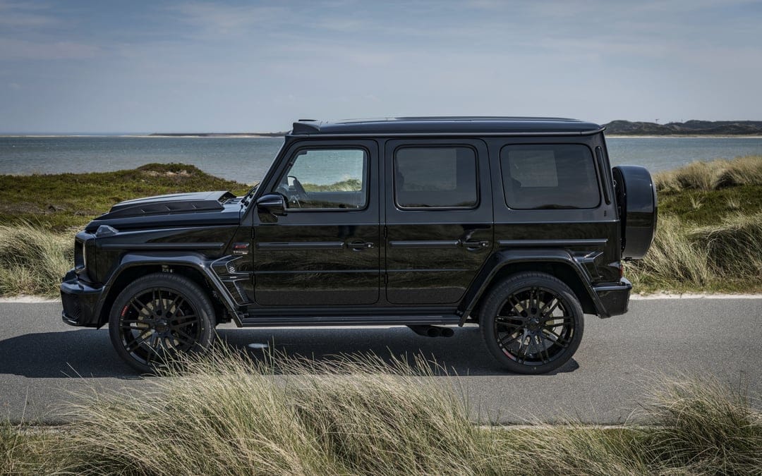Black Brabus G-Class SUV parked on a coastal road beside grassy dunes under a clear blue sky with ocean horizon.