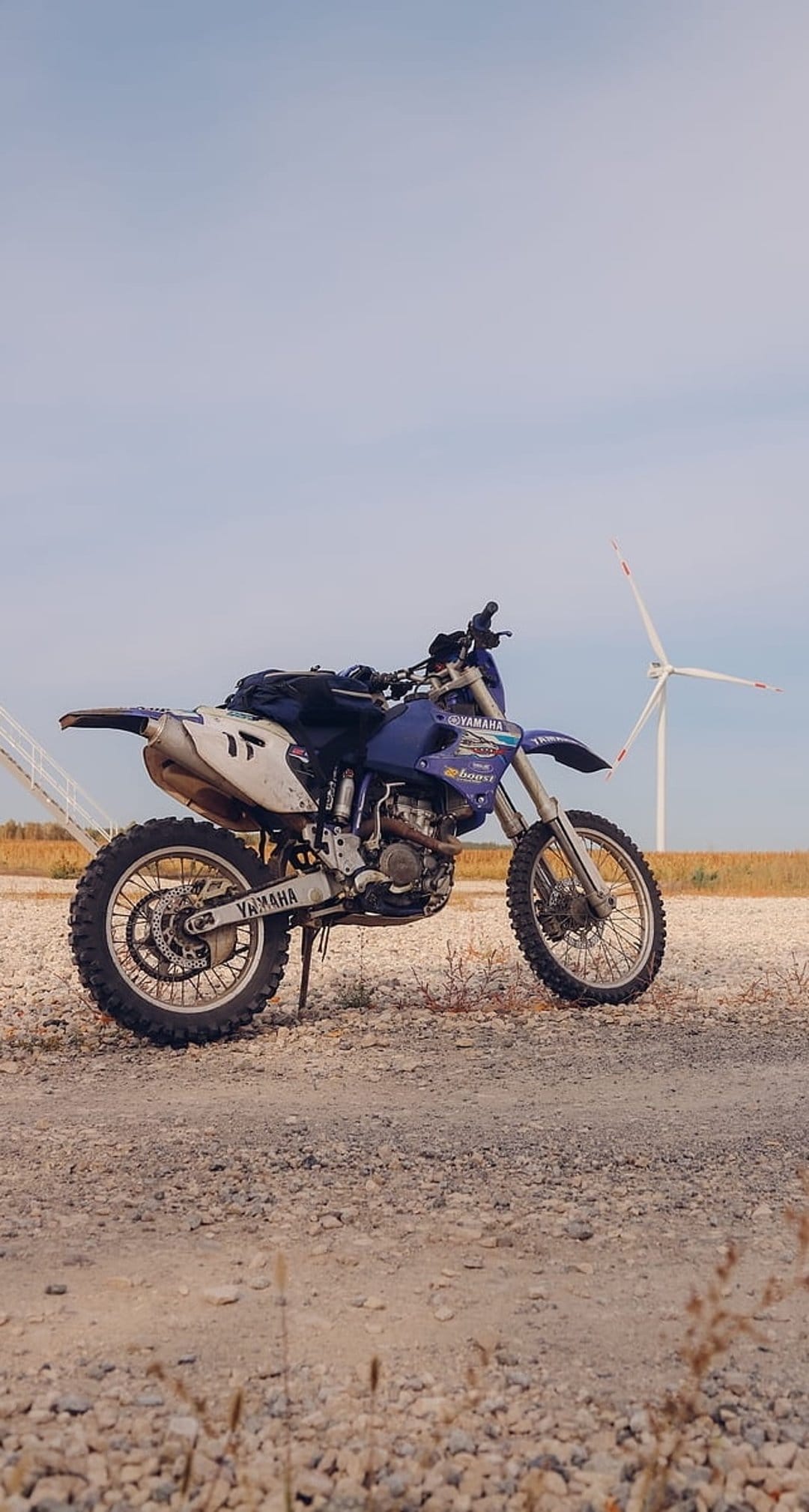 Blue Yamaha dirt bike parked on a gravel road with a white wind turbine under a cloudy sky in a rural field.