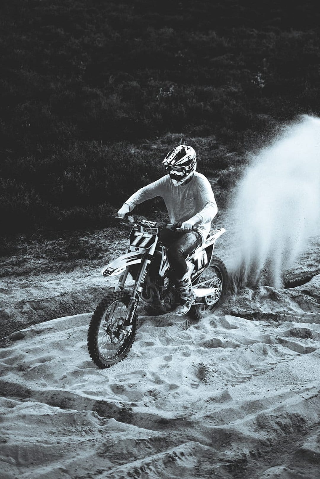 Motocross rider wearing a helmet leans a dirt bike into a sandy turn, kicking up a large plume of dust in monochrome.