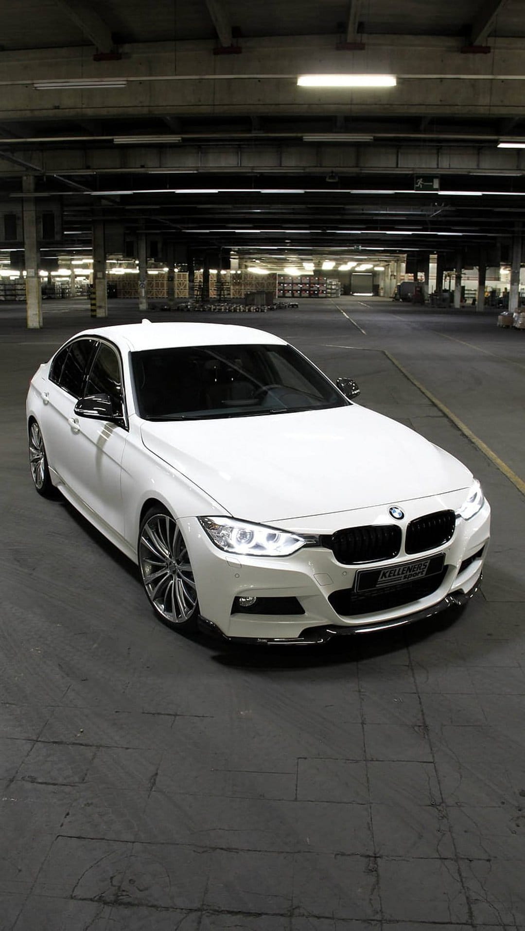 White BMW M Sport sedan with tinted windows parked on a concrete floor in a dark industrial warehouse garage.