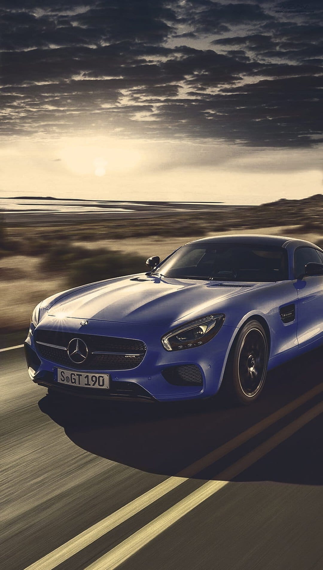 Blue Mercedes-AMG GT S sports car speeds on a road under a dramatic sunset sky with motion blur and golden light.