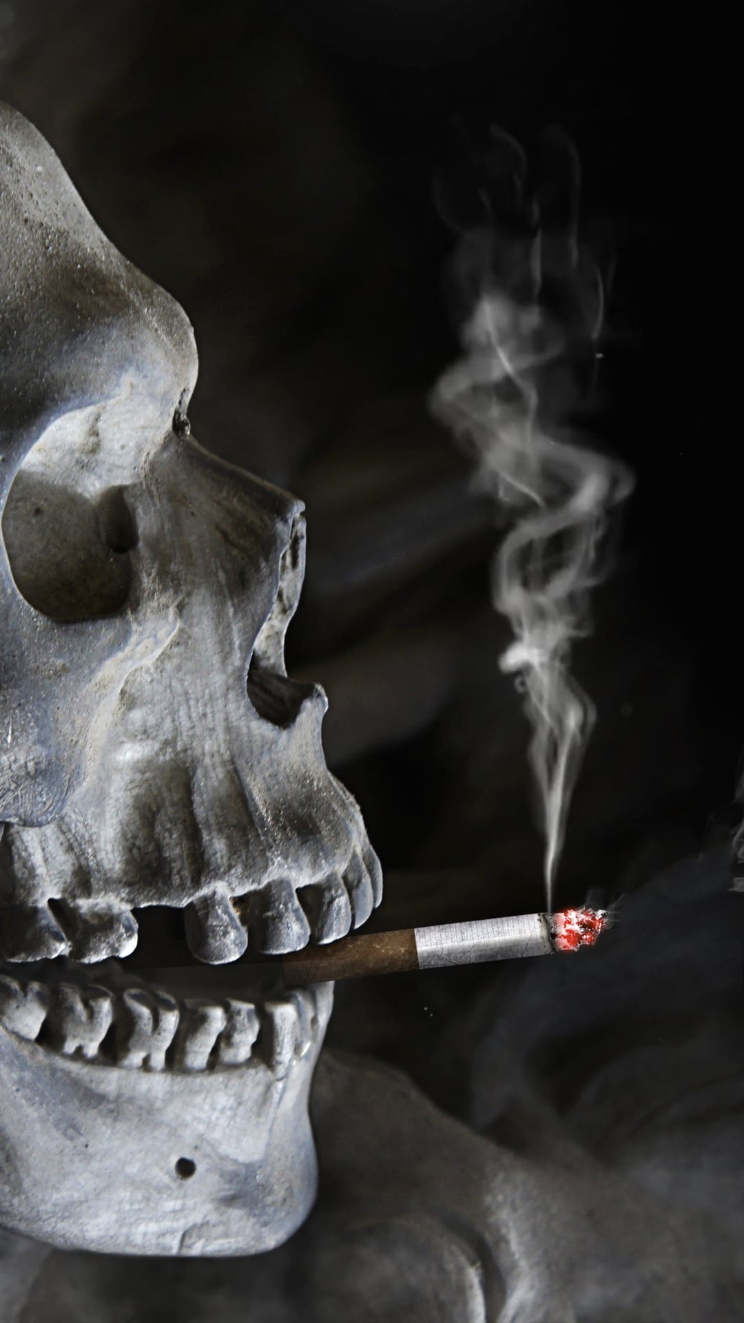 Stone skull holding a lit cigar with rising wisps of smoke and a glowing ember against a black background.