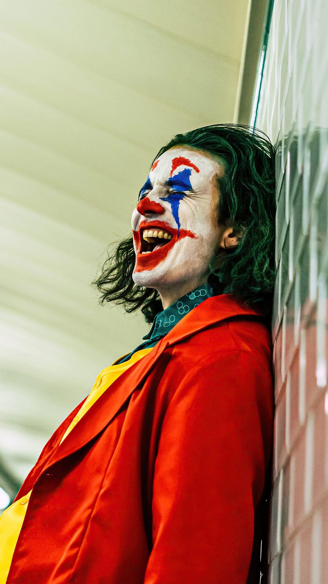 Arthur Fleck as Joker laughing against a white wall wearing a red coat and colorful clown face paint.