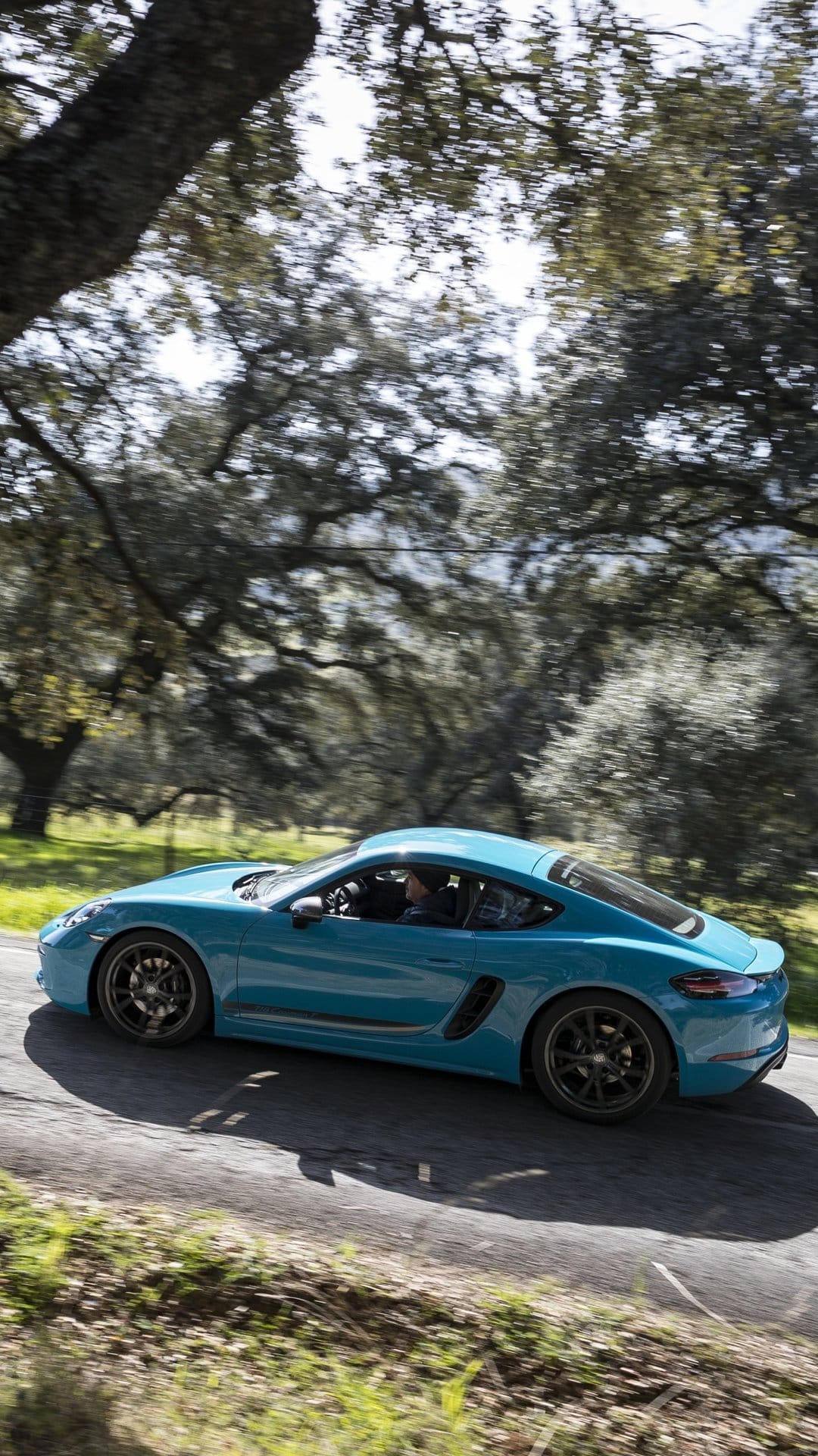 Blue Porsche 718 Cayman T driving fast on a forest road with motion blurred trees and sun-dappled lighting.